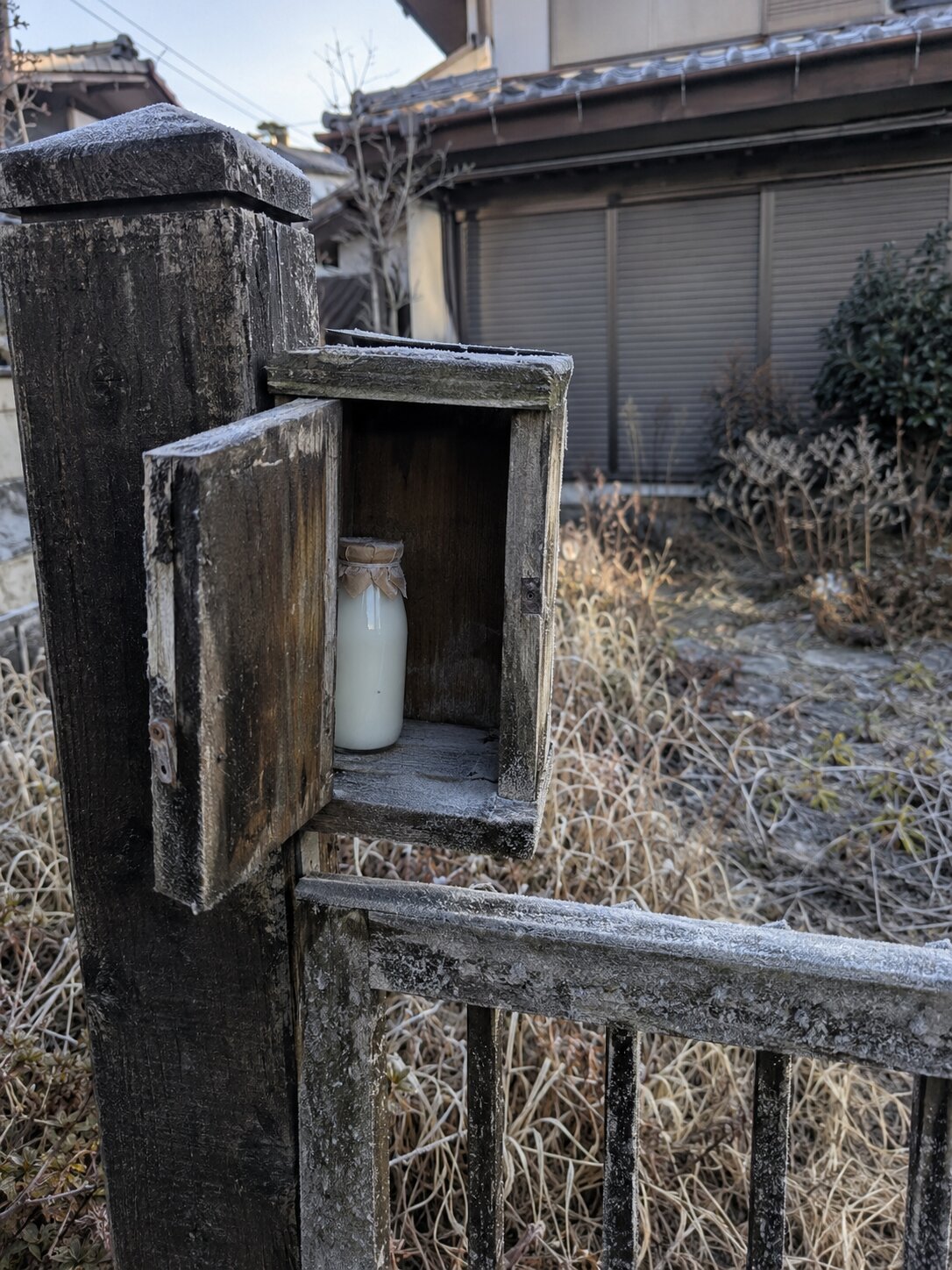向かいの家の女性撮影。霜のついた古い木の牛乳箱の扉が開いていて、中にガラスの牛乳瓶が1本入っている。