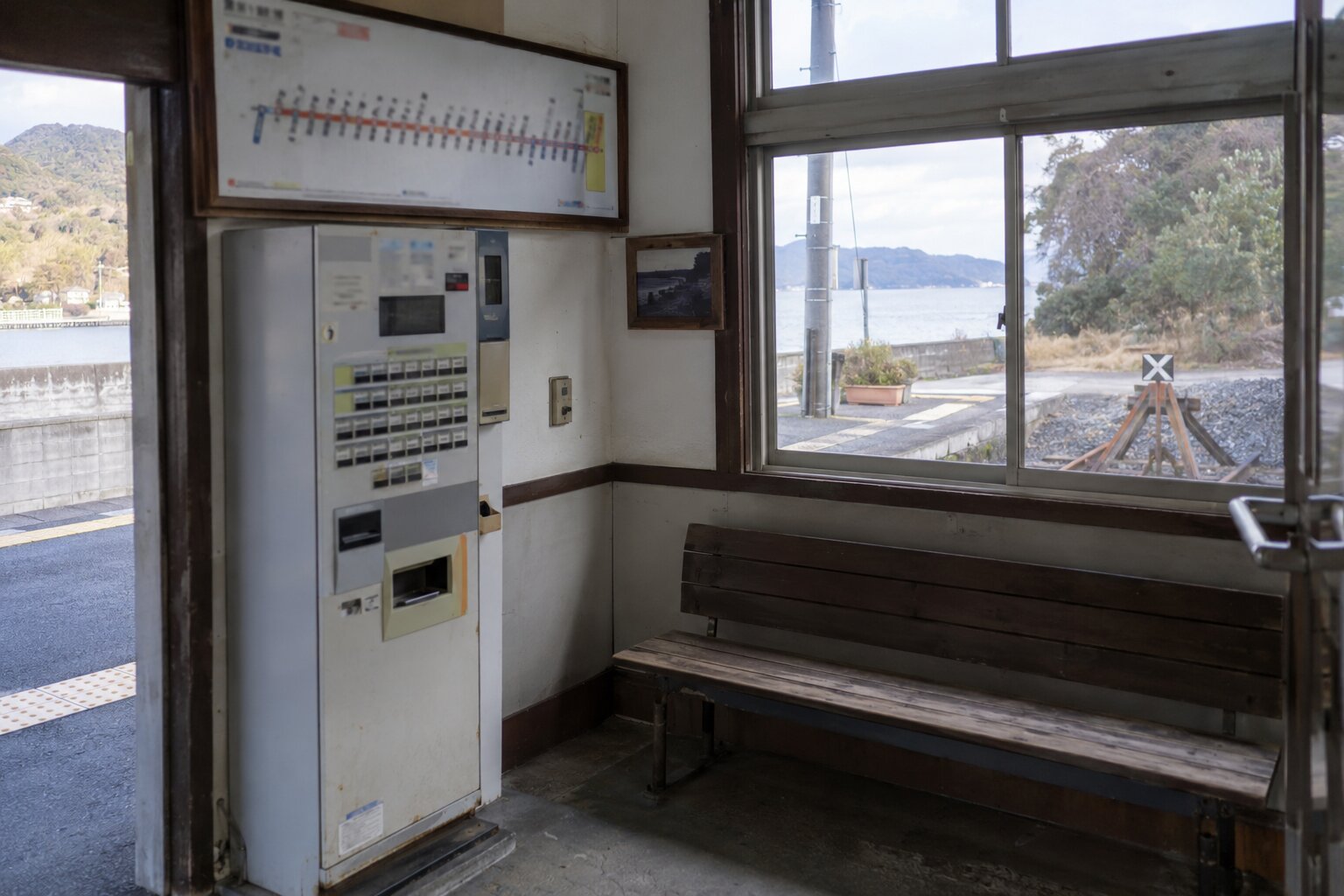 霧島秀一撮影。小さな駅舎の中の古い券売機と木のベンチ。窓の外に海と線路の車止めが見える。