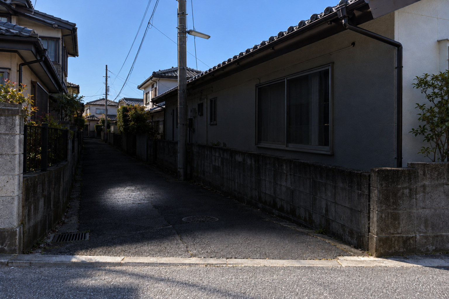霧島秀一撮影。2023年3月28日午前。青空と日を受ける屋根、明るい手前の通りの奥に、暗い路地が続く。街灯は消えており、懐中電灯の丸い光が当たる舗装の小石とひびだけが明るく見える。