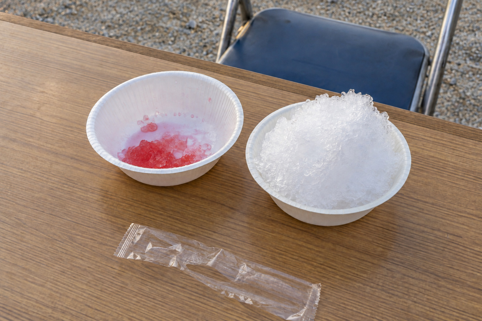 霧島秀一撮影。長机の上に白い紙椀が2つ並び、片方の底には赤い氷が少し残り、隣の椀には白い氷が盛られている。開封したスプーンの袋が手前にある。