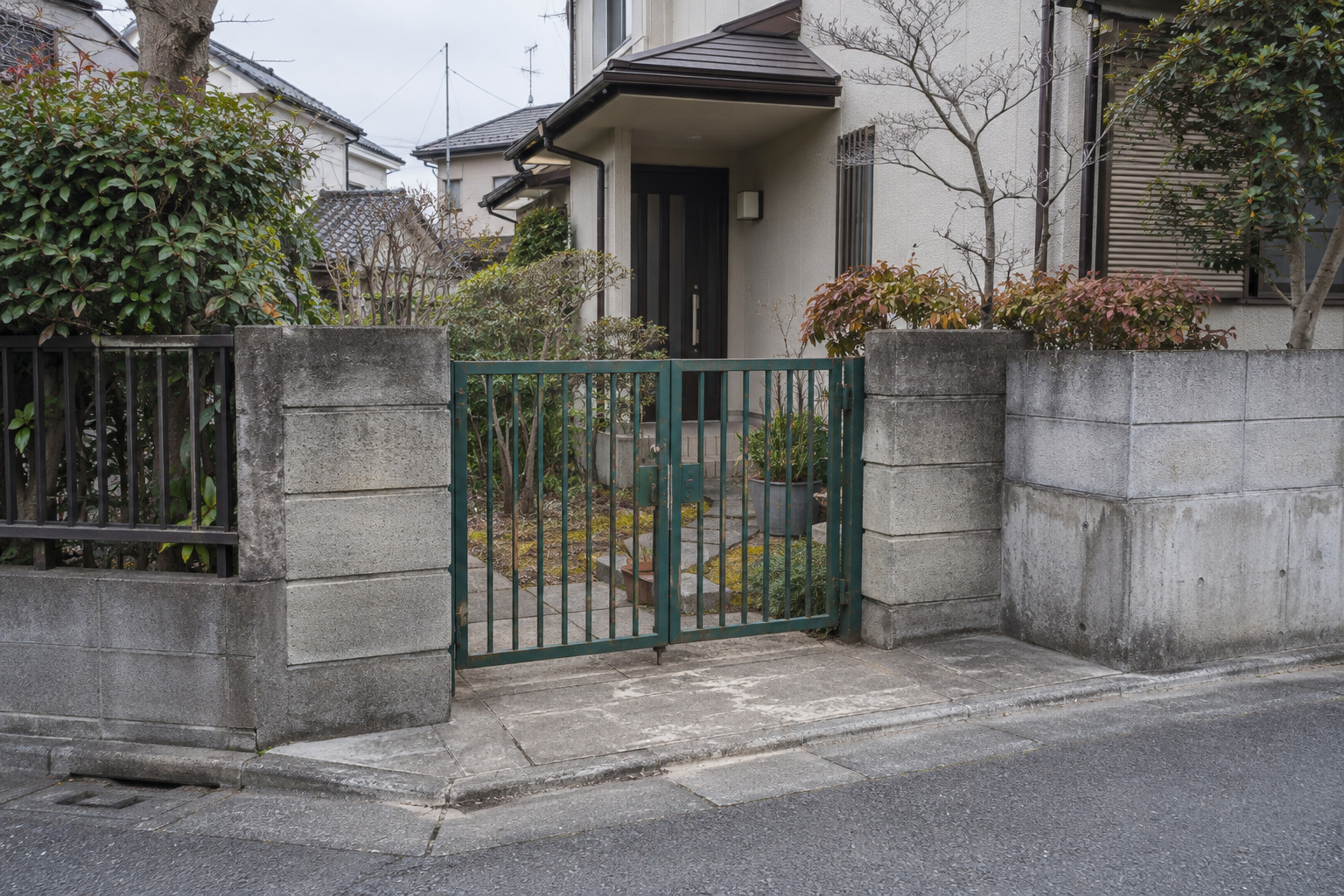 霧島秀一撮影。住宅の小さな前庭と、道路に面した緑色の鉄の門。門は閉じていて、門柱の外側には人が座れる舗装面がある。