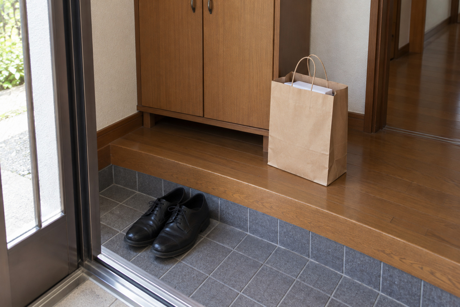 霧島秀一撮影。玄関の上がり口の脇に置かれた茶色の紙袋と、脱いだ黒い靴。紙袋の口から、白い包装紙に包まれた四角い菓子箱が見えている。