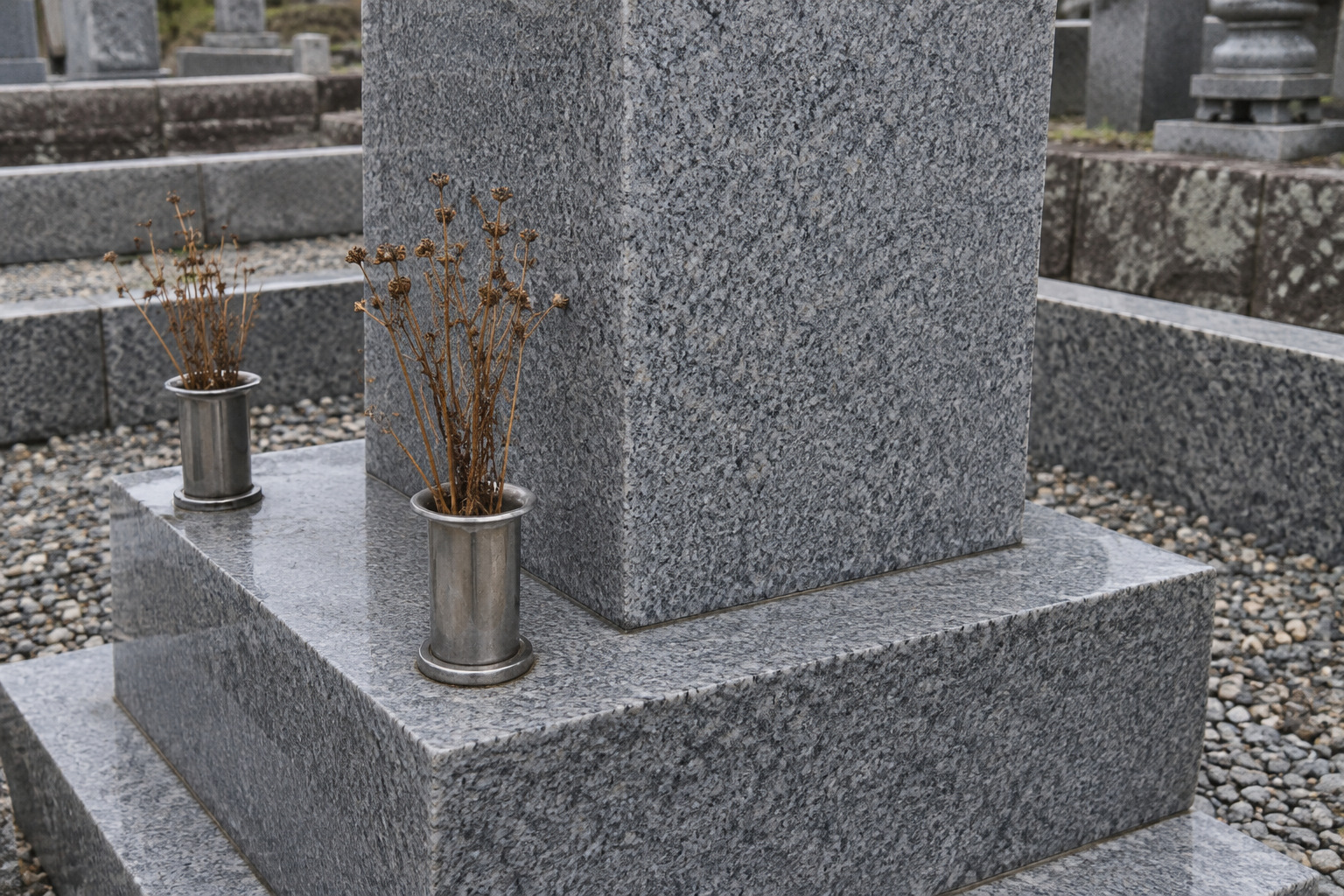 霧島秀一撮影。灰色の四角い墓石の平らな側面と、その手前の金属の花立て。側面に穴や割れ目は見えず、花立てには枯れた茎が残っている。